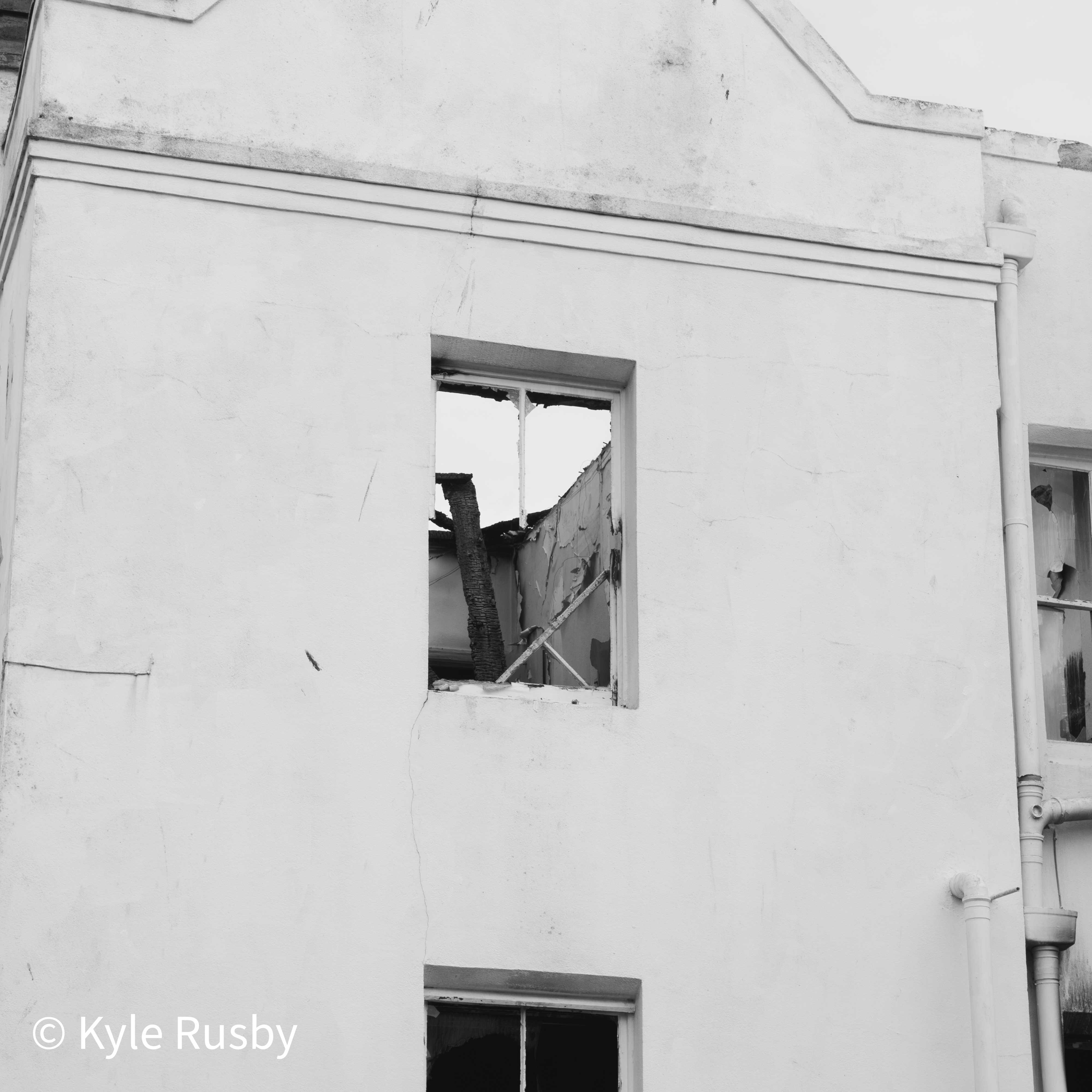 Black and white photograph of a fire-gutted house, charred roof timbers visible through an empty upstairs window in a cracked white gable