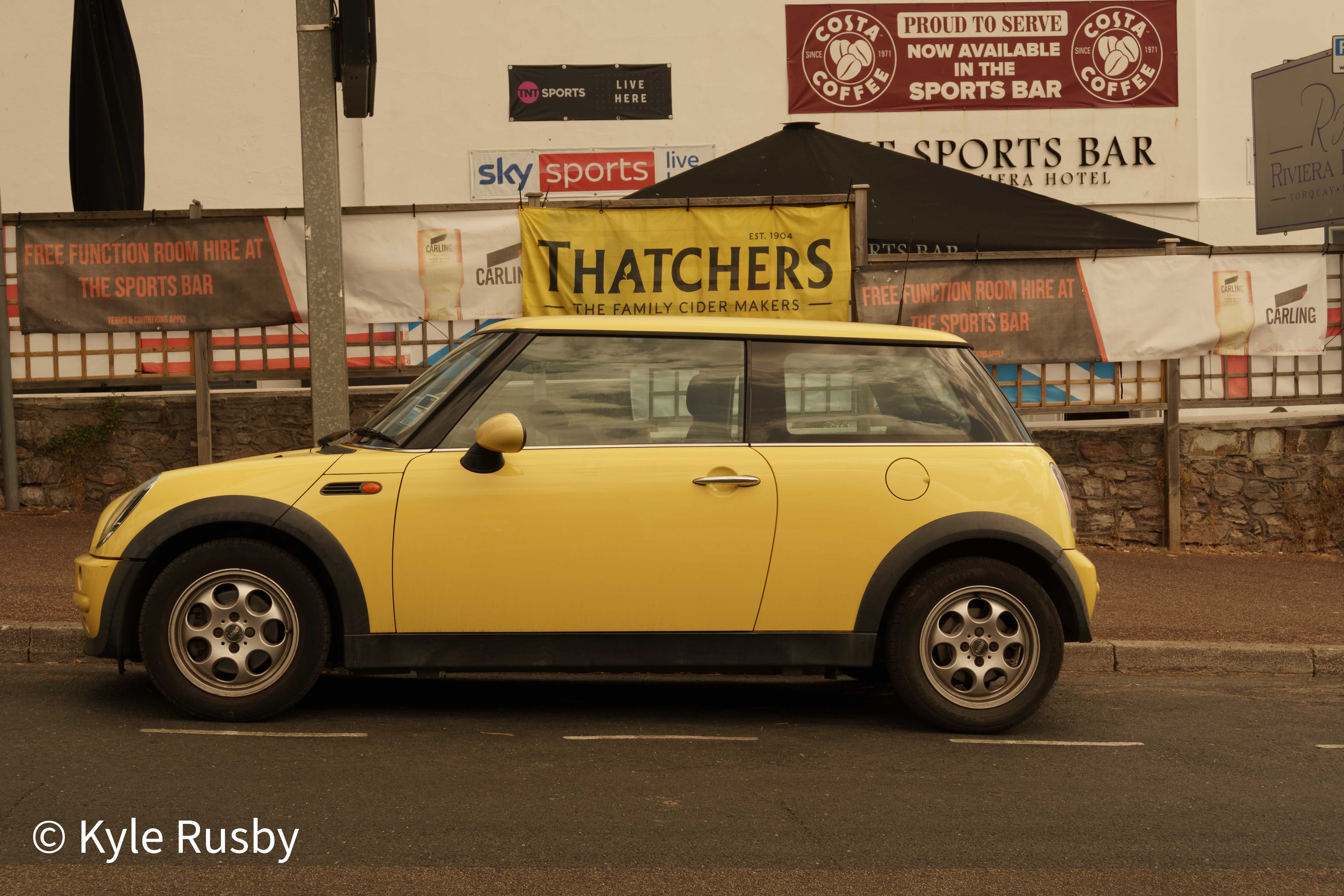 A yellow Mini parked at the kerb beneath a Thatchers cider banner, sports bar signs and hazy pink light behind