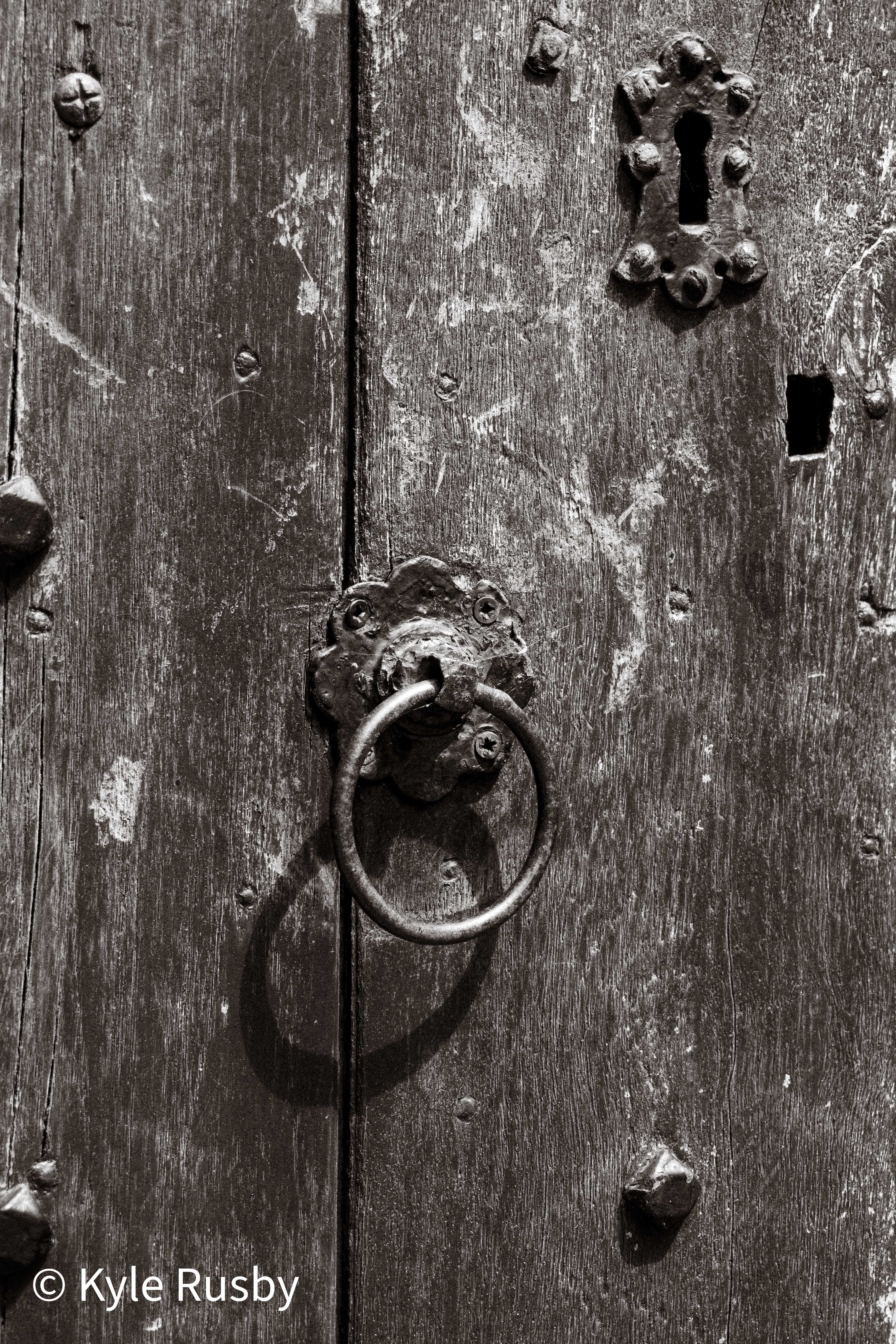 Black and white study of a scarred medieval door: an iron ring handle on a flower-shaped plate below an ornate keyhole escutcheon