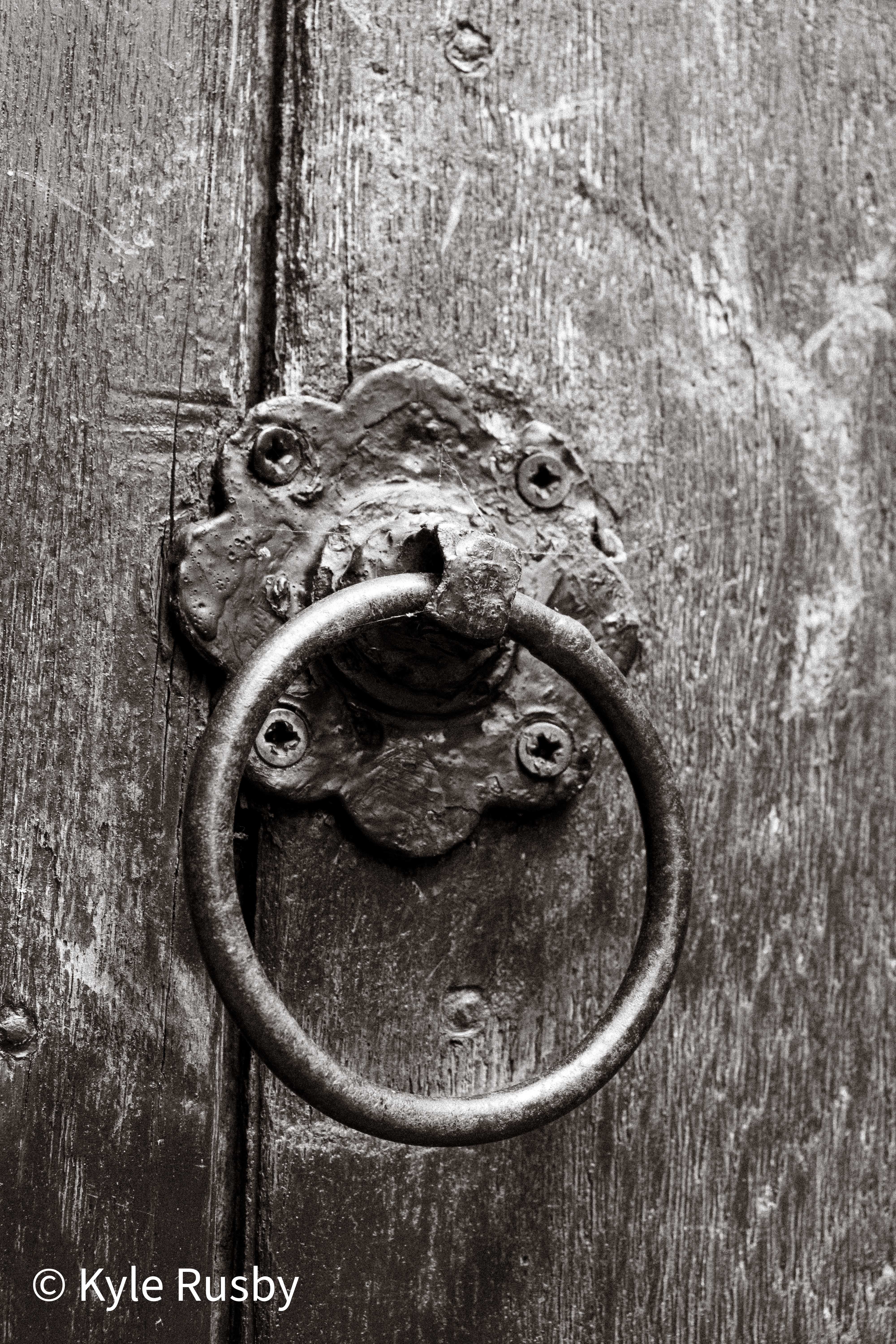 Close black and white detail of the same door: an iron ring hanging from a flower-shaped backplate on deeply grained wood