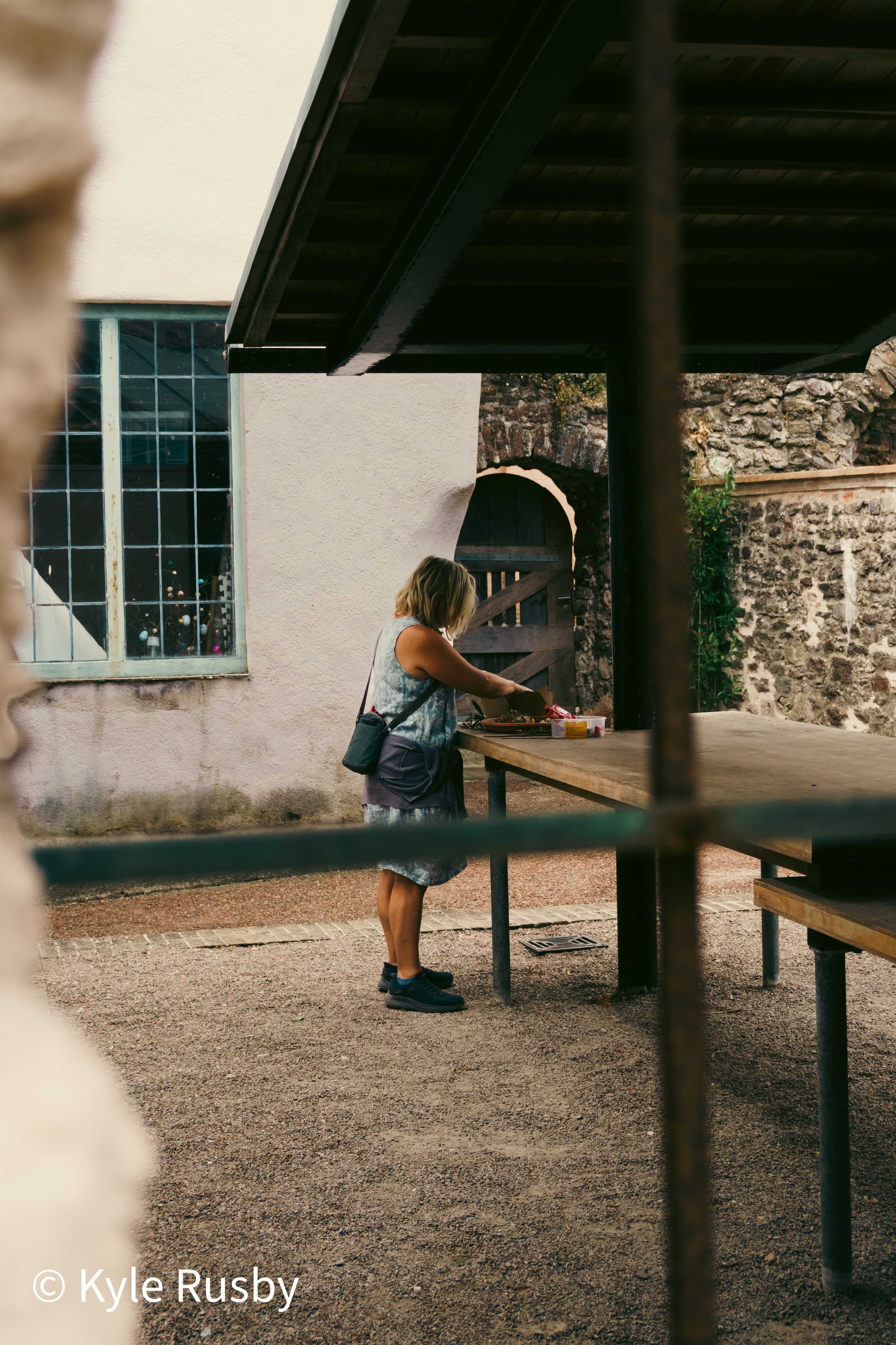Seen between railings, a woman lays out lunch at a long table under a dark canopy in a stone courtyard, an arched wooden door behind