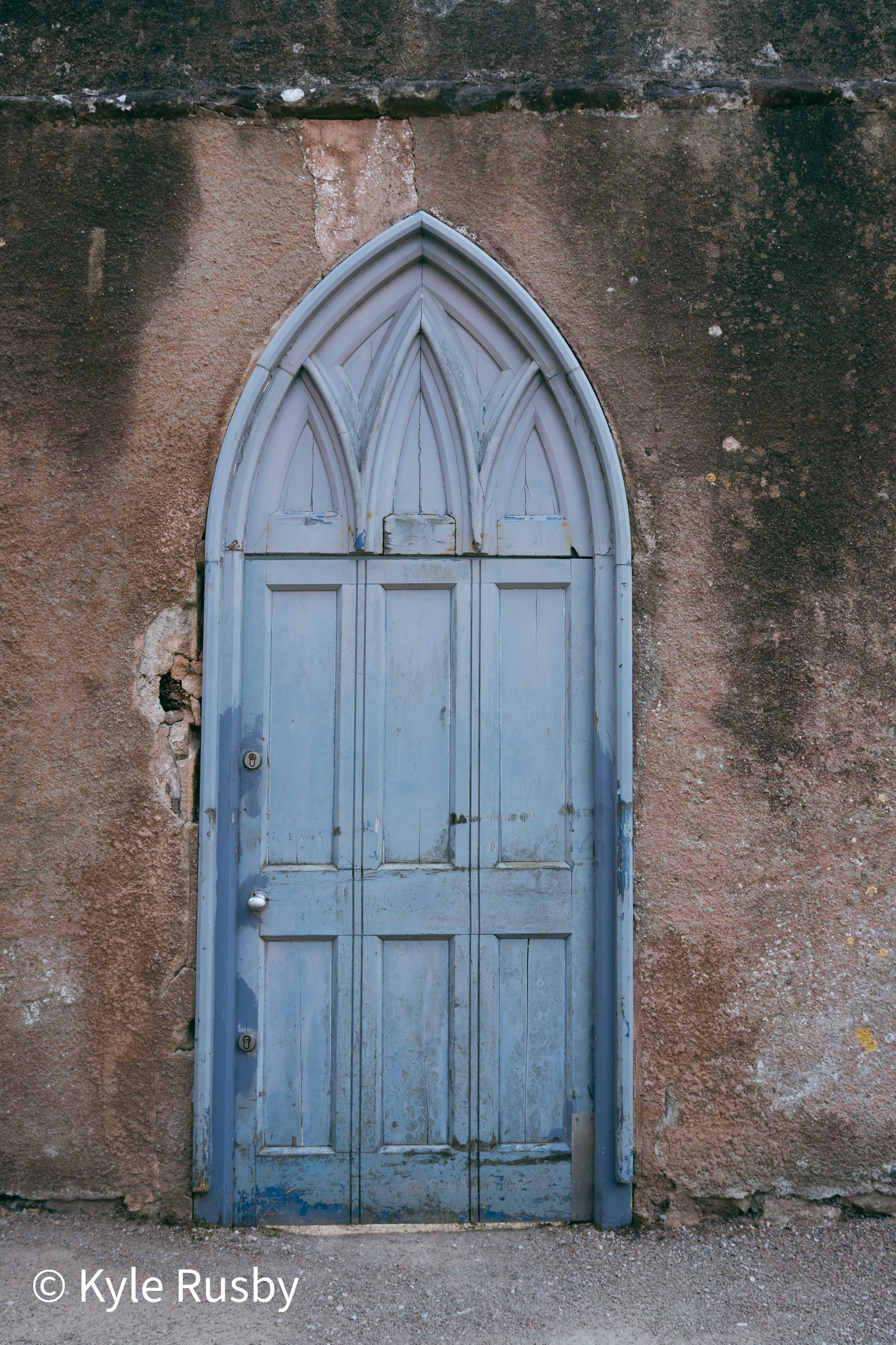 A powder-blue gothic arched door with peeling panels set into a weathered wall of pink and ochre render