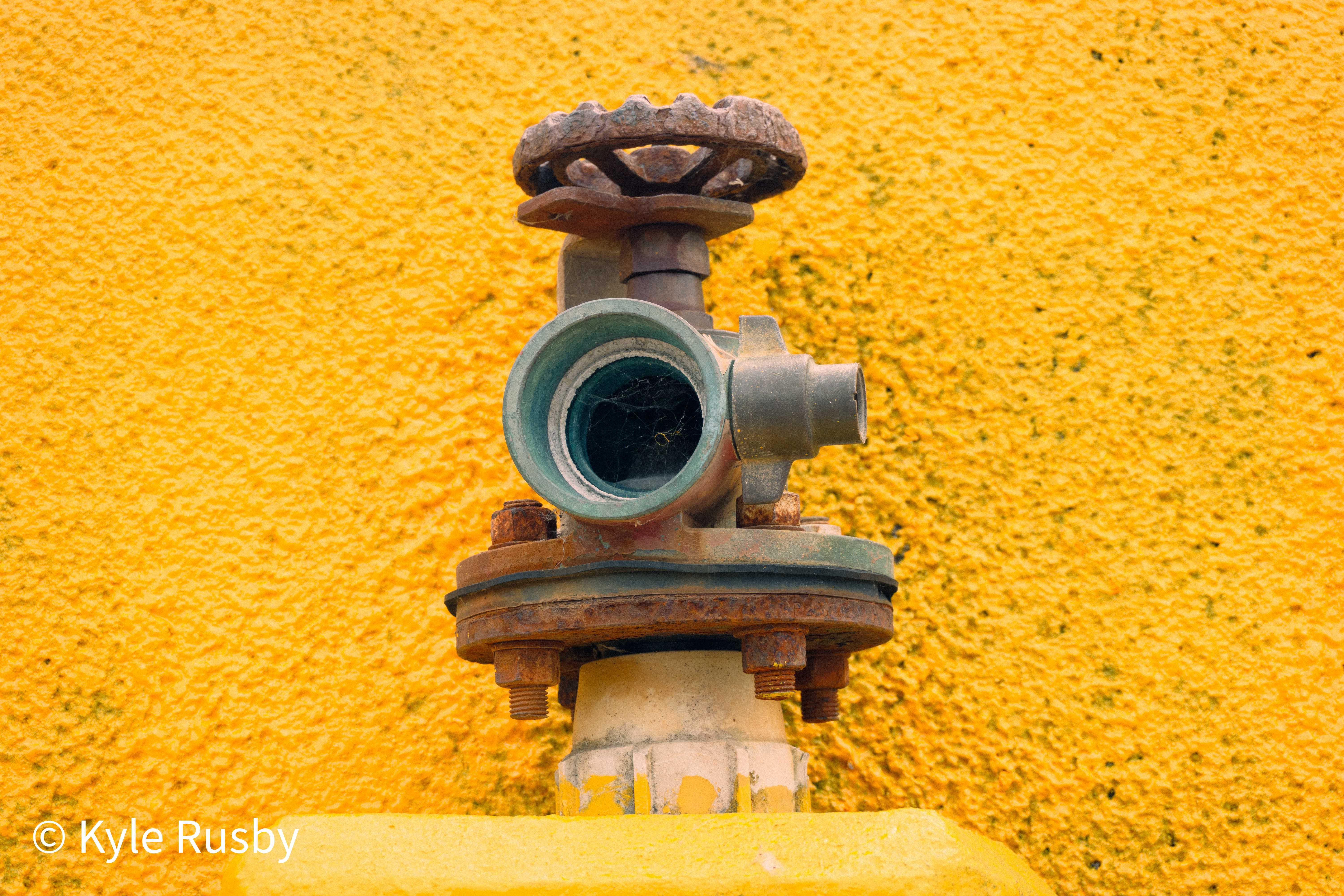 An old verdigris water valve with a rusted handwheel bolted to a wall of bright yellow pebbledash