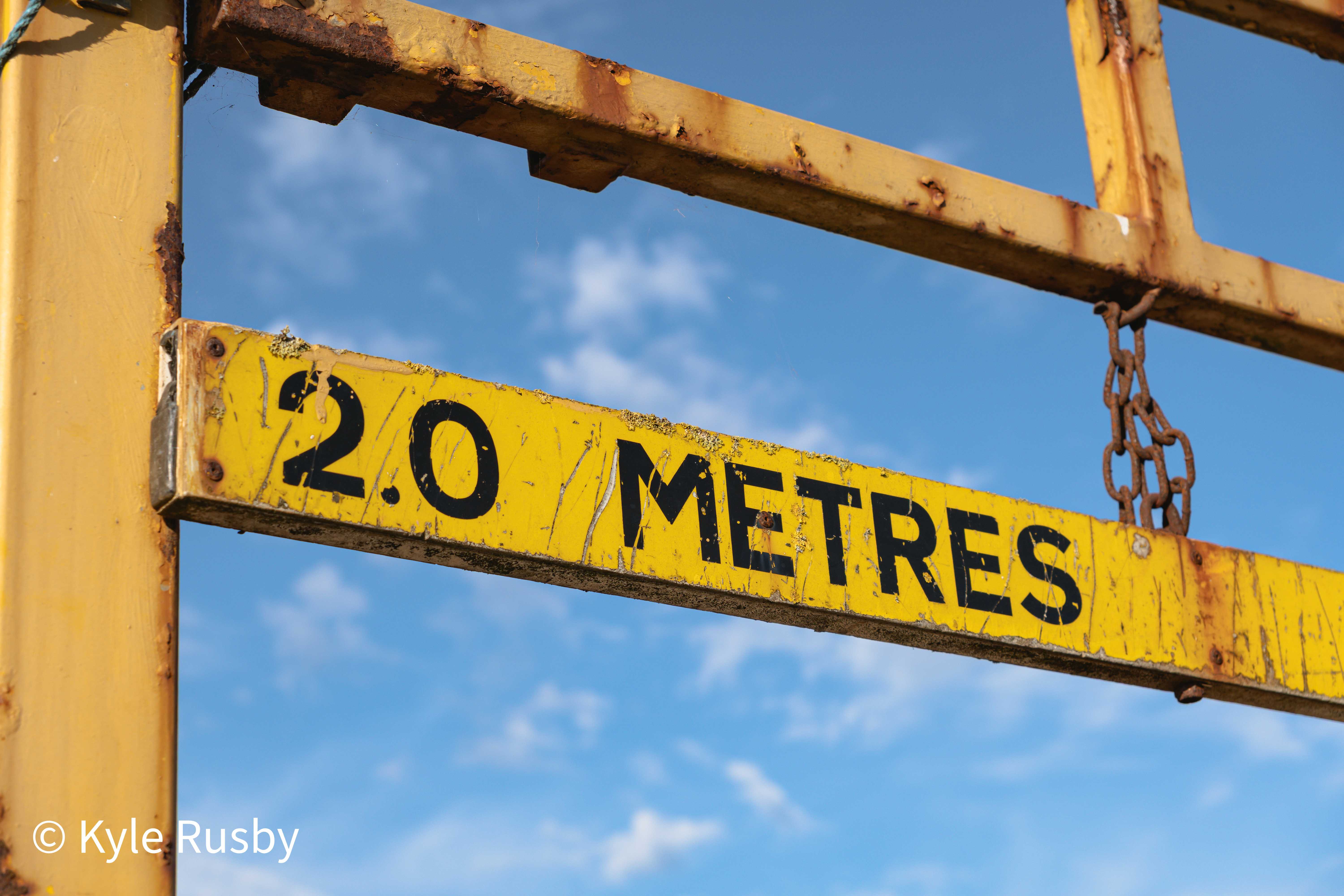 A rusting yellow height barrier lettered 2.0 METRES hangs on its chain against a blue sky of small clouds