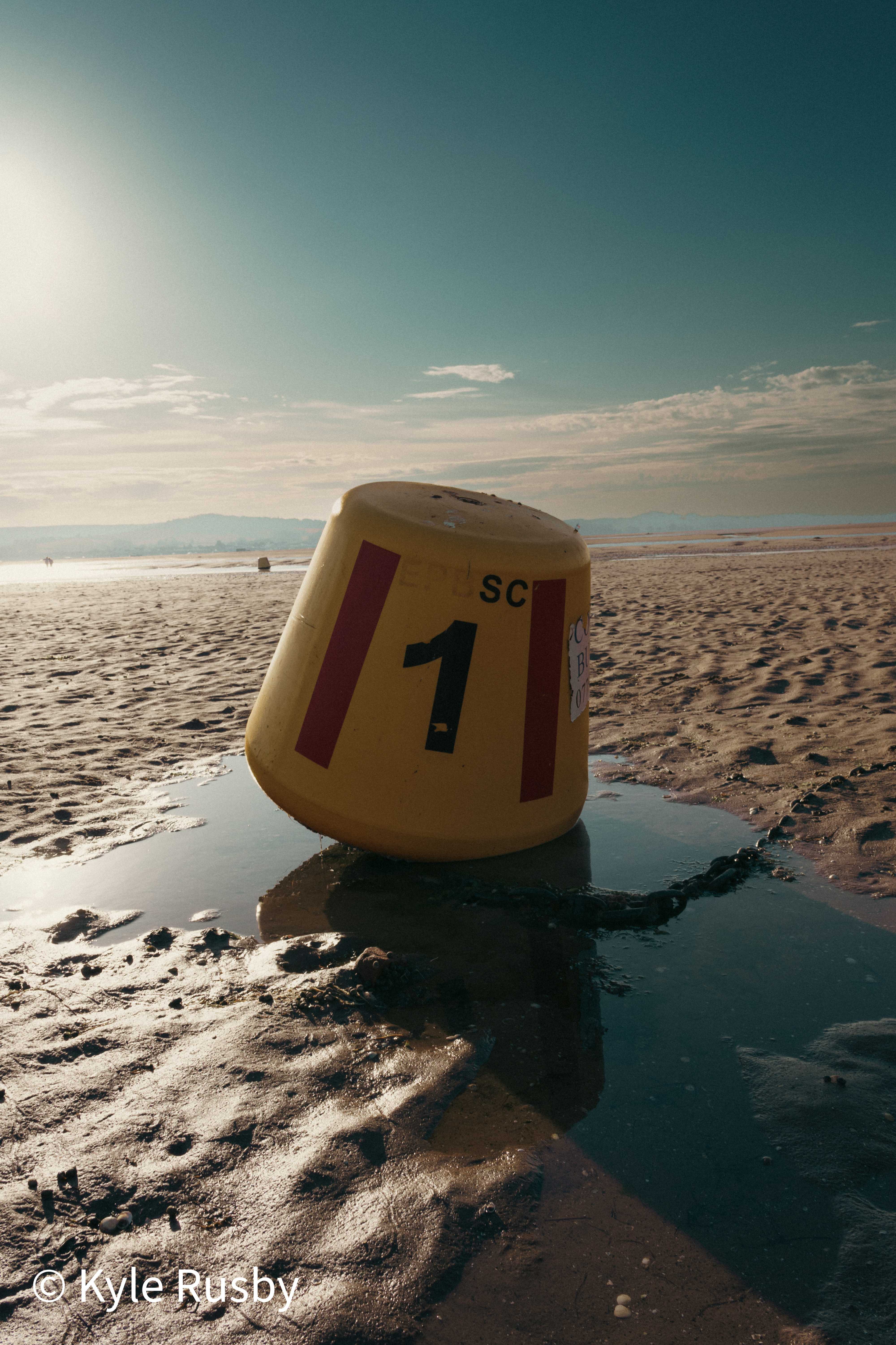 A yellow buoy marked with a large number 1 sits aground on rippled wet sand, its chain trailing into a tide pool against the low sun