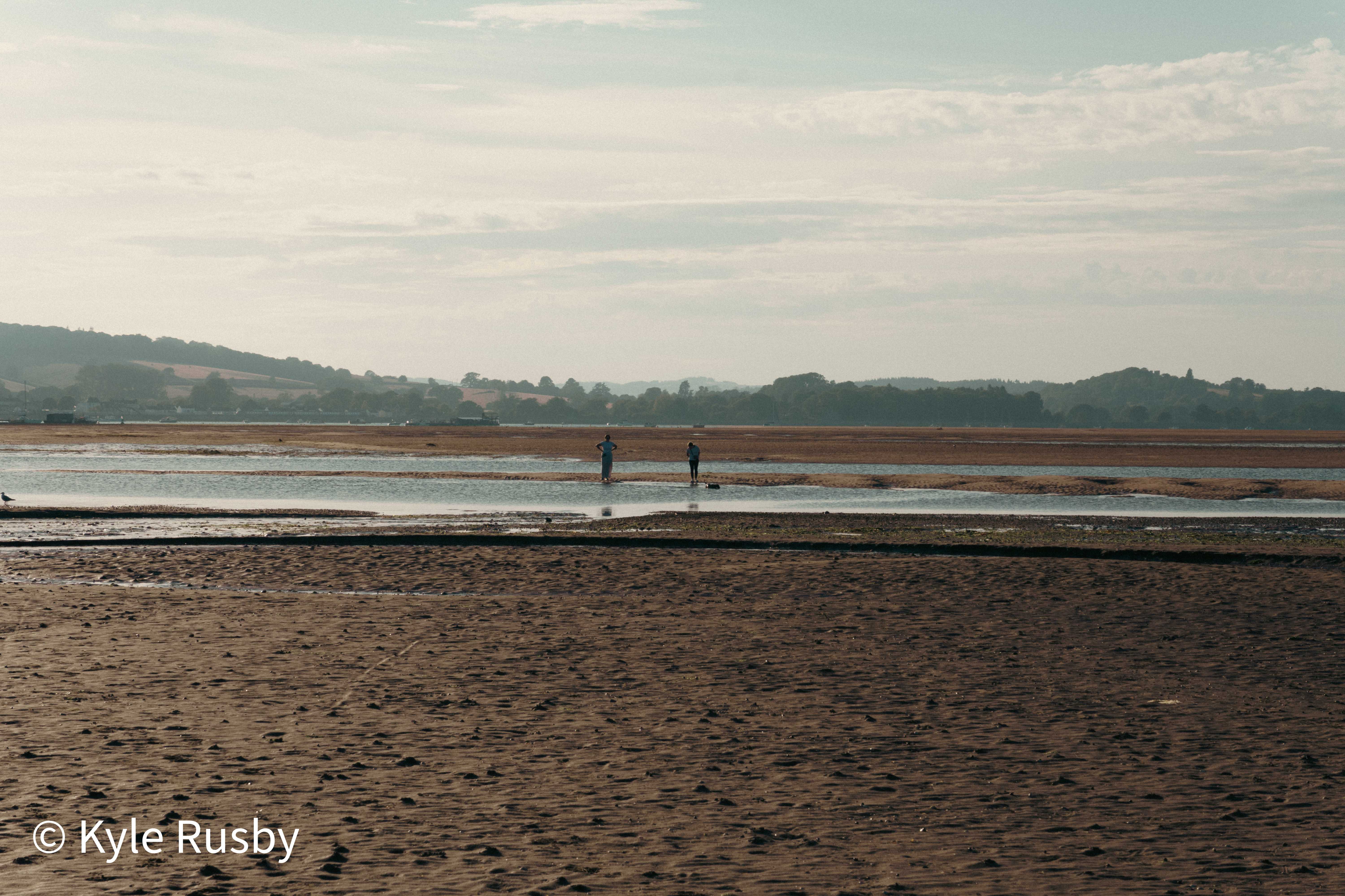 Two small figures wade the last channel of water left in the sands, the far hills pale in the morning haze