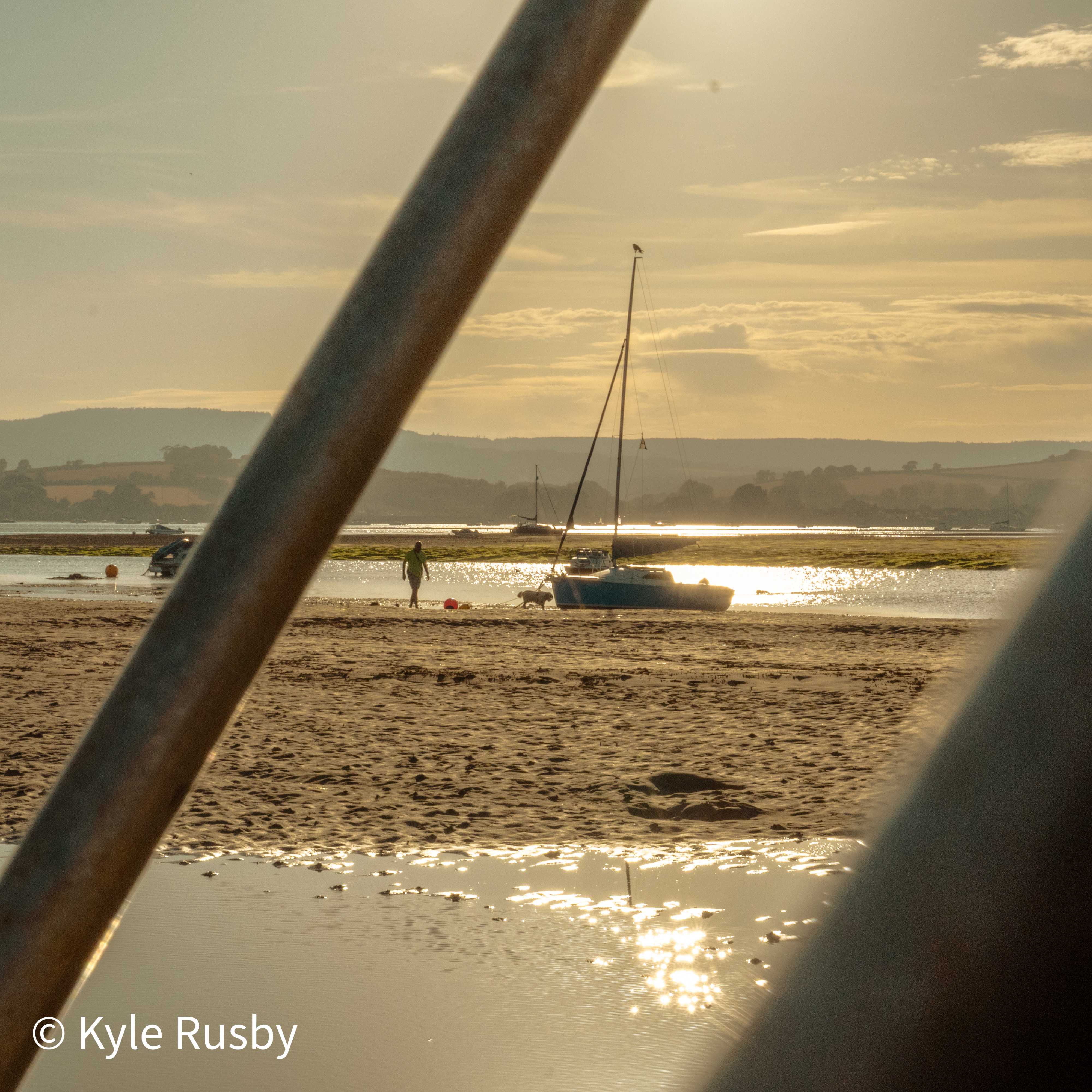 Seen between two blurred railings, a blue sailboat sits aground at golden hour while a figure and dog cross the sand