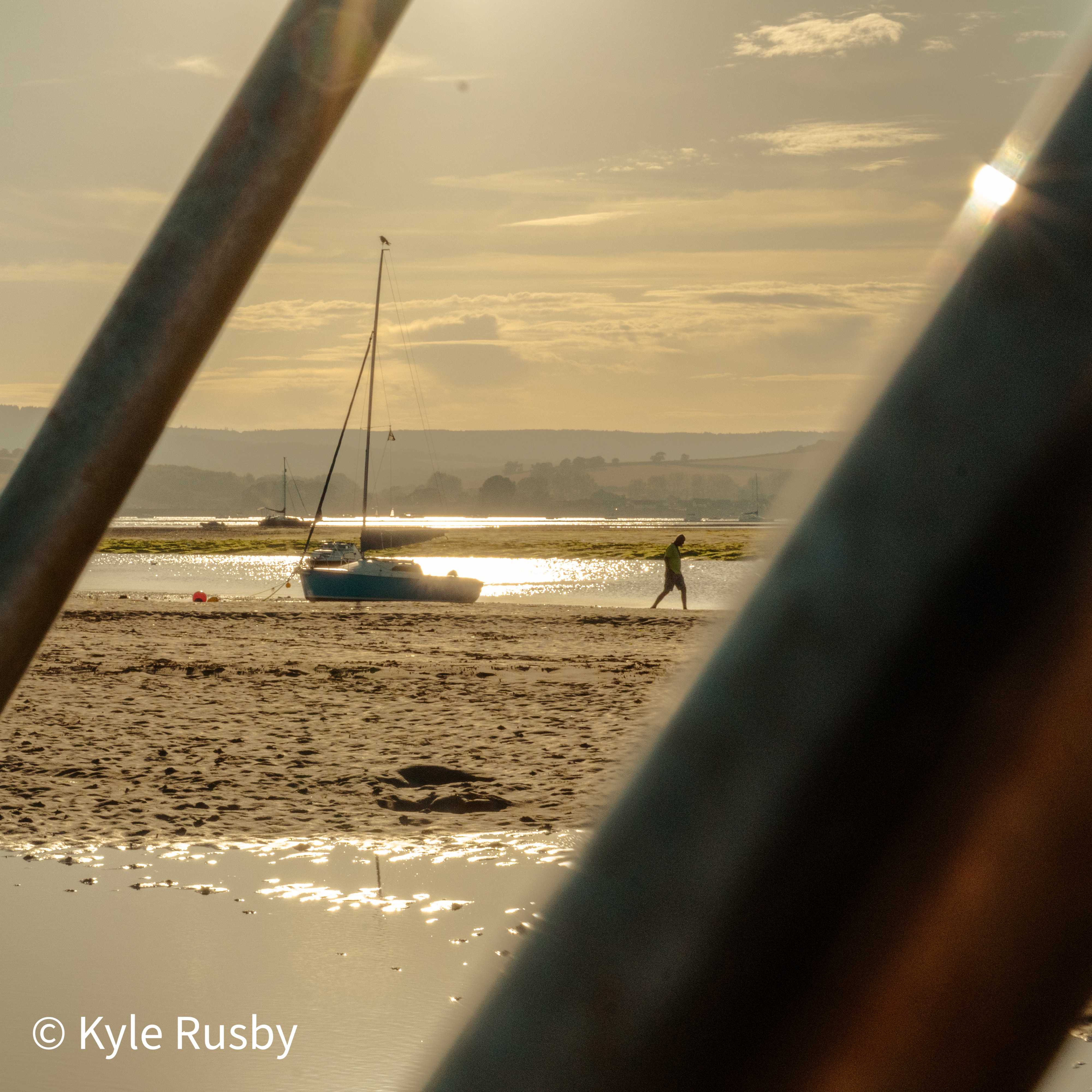 The blue sailboat aground on glittering wet sand between two railings, a figure in a high-vis jacket walking away along the water