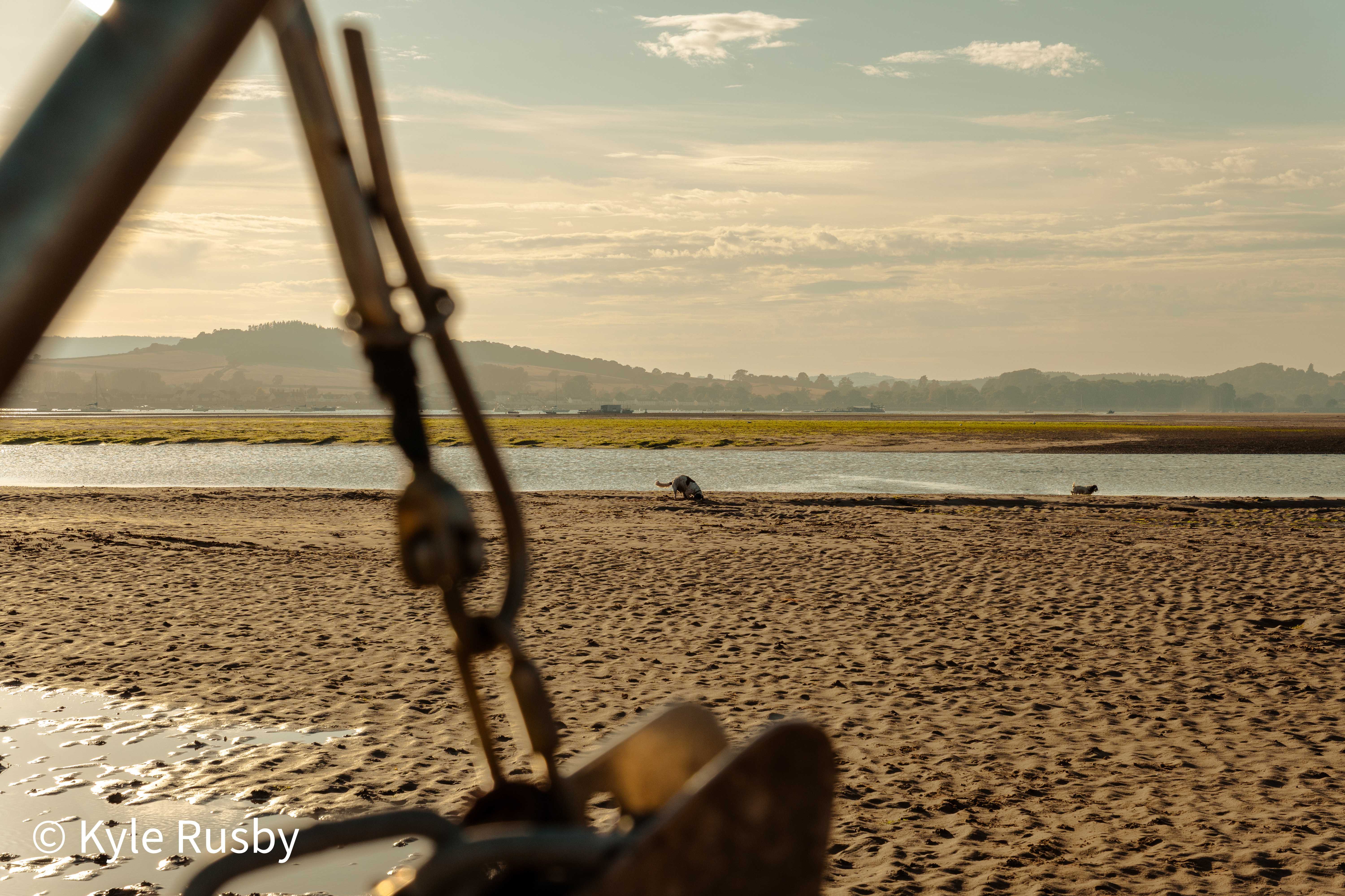 A blurred anchor and rigging in the foreground frame the emptied estuary, a distant dog splashing through the shallows beyond