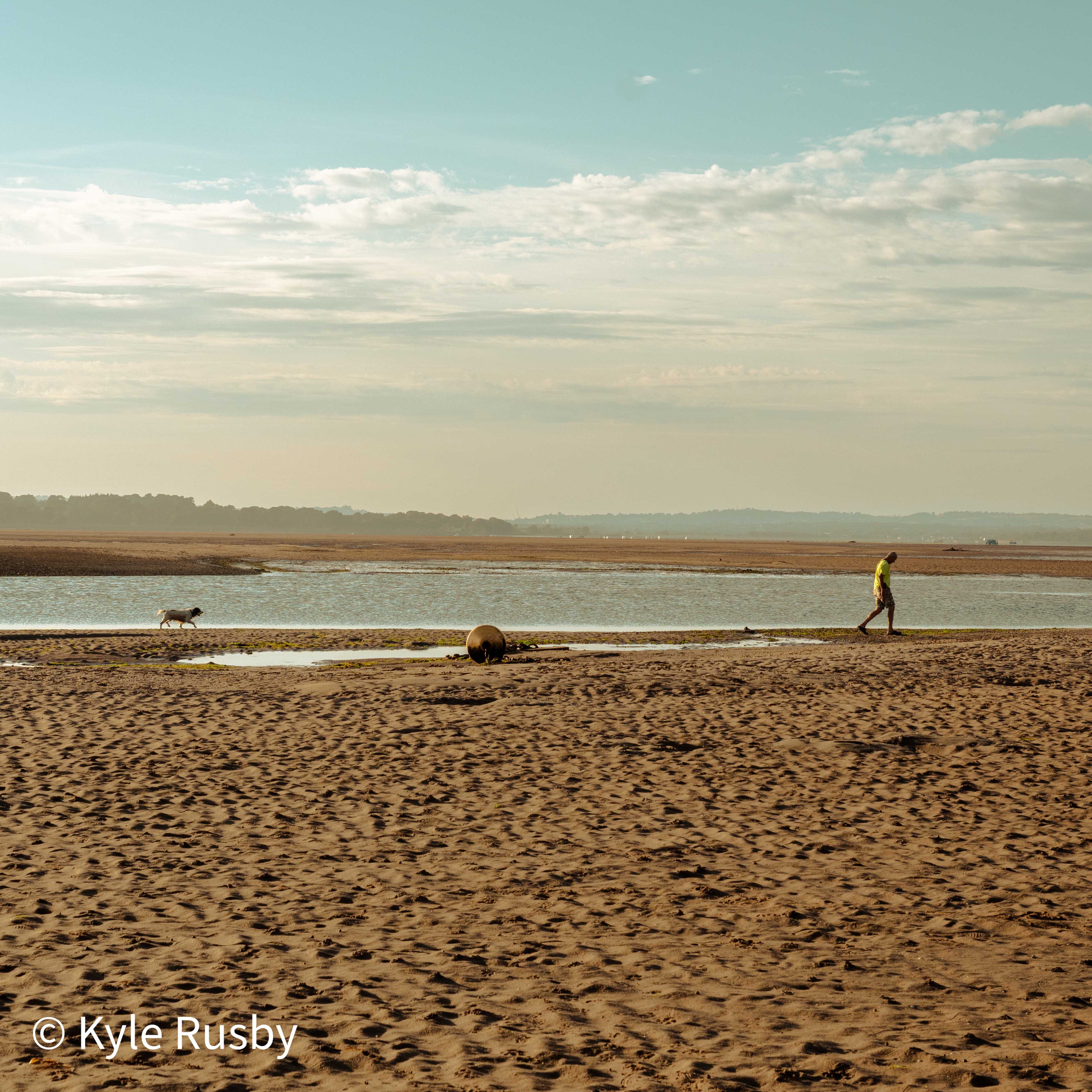 A walker in yellow and their dog follow the edge of a tide pool across wide rippled sand under a big morning sky