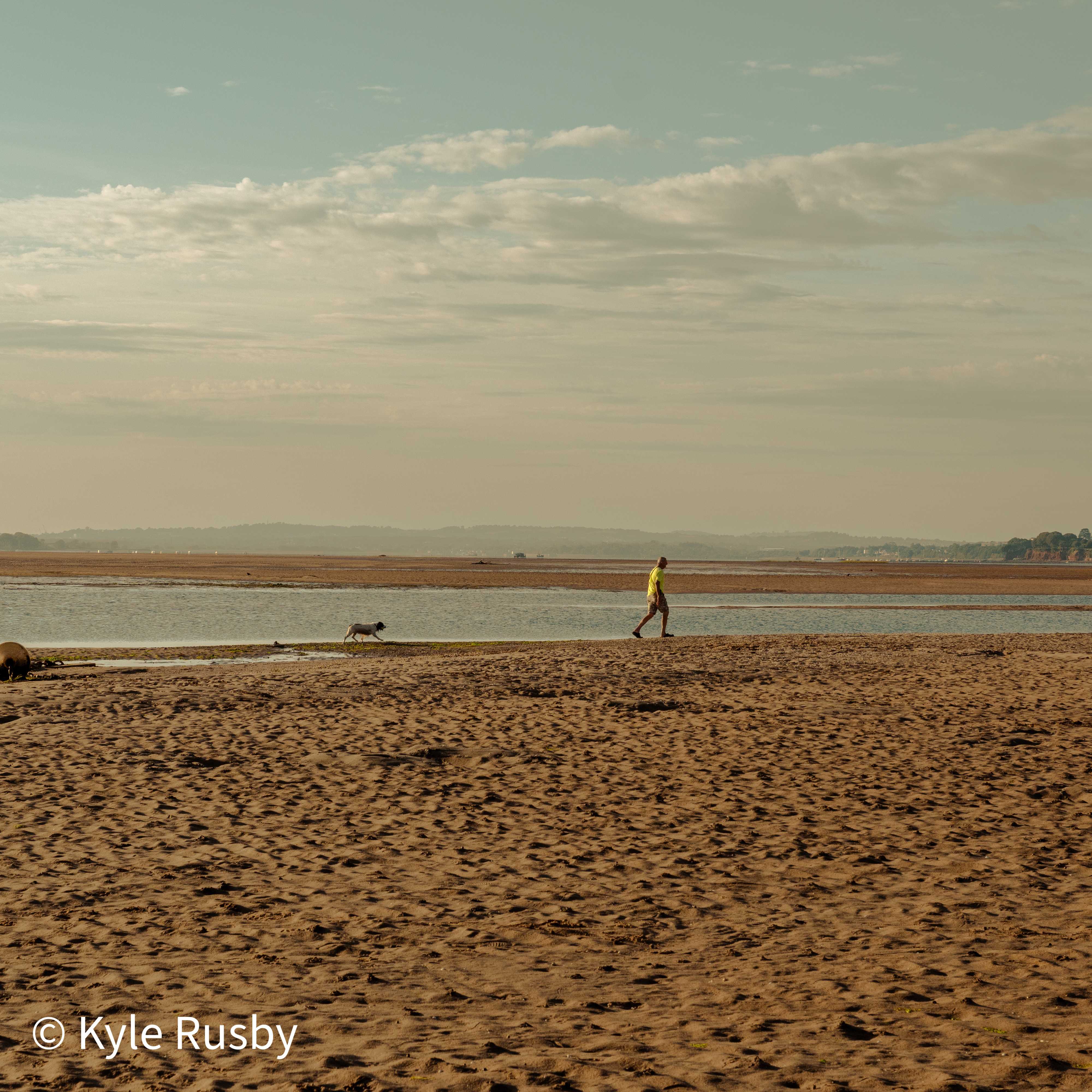 The same walker striding the sandbar mid-step, the dog trotting the waterline ahead of them