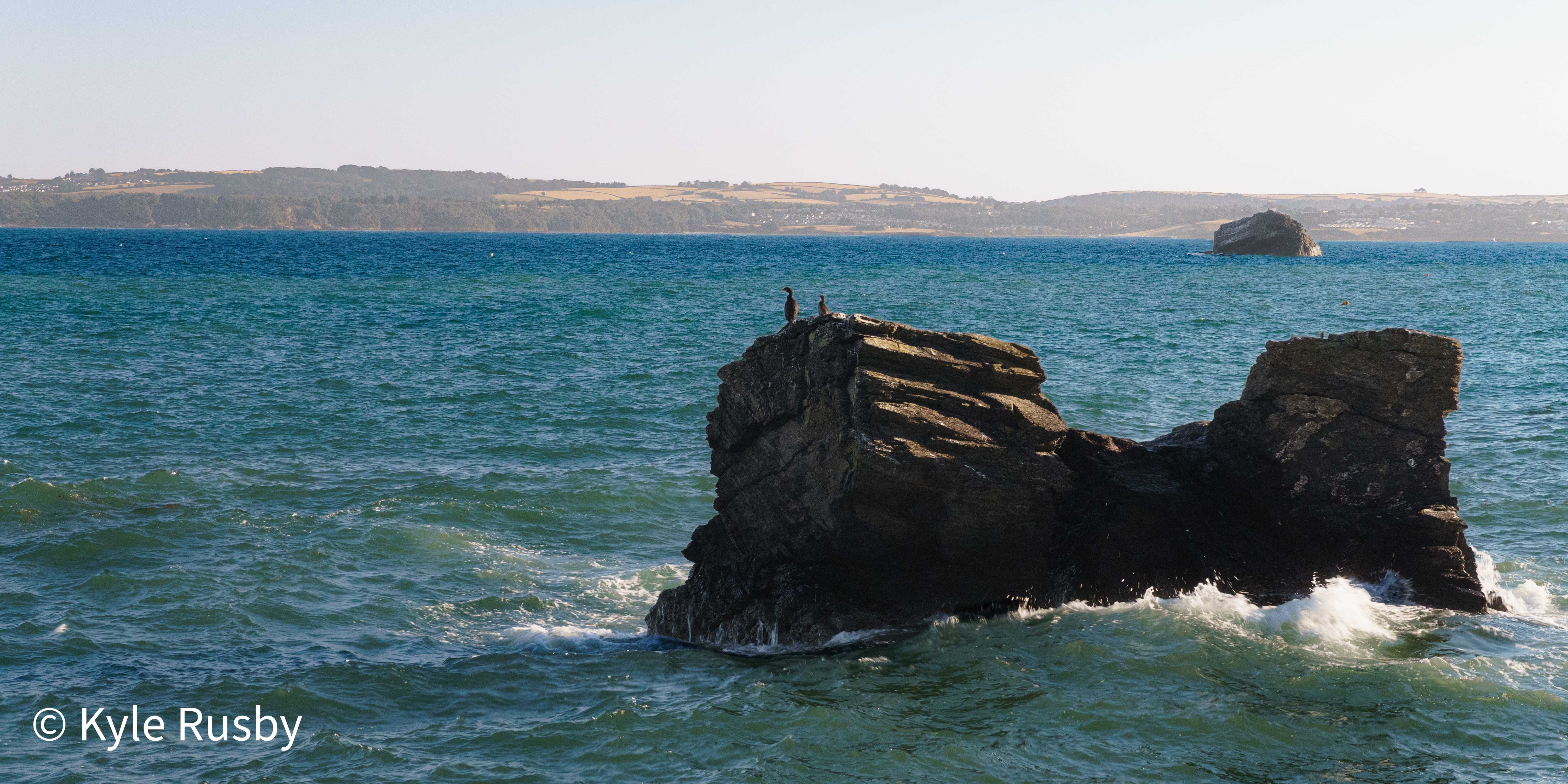 Two cormorants stand on a layered rock stack in a choppy teal sea, waves breaking white at its base and the far coast hazy behind