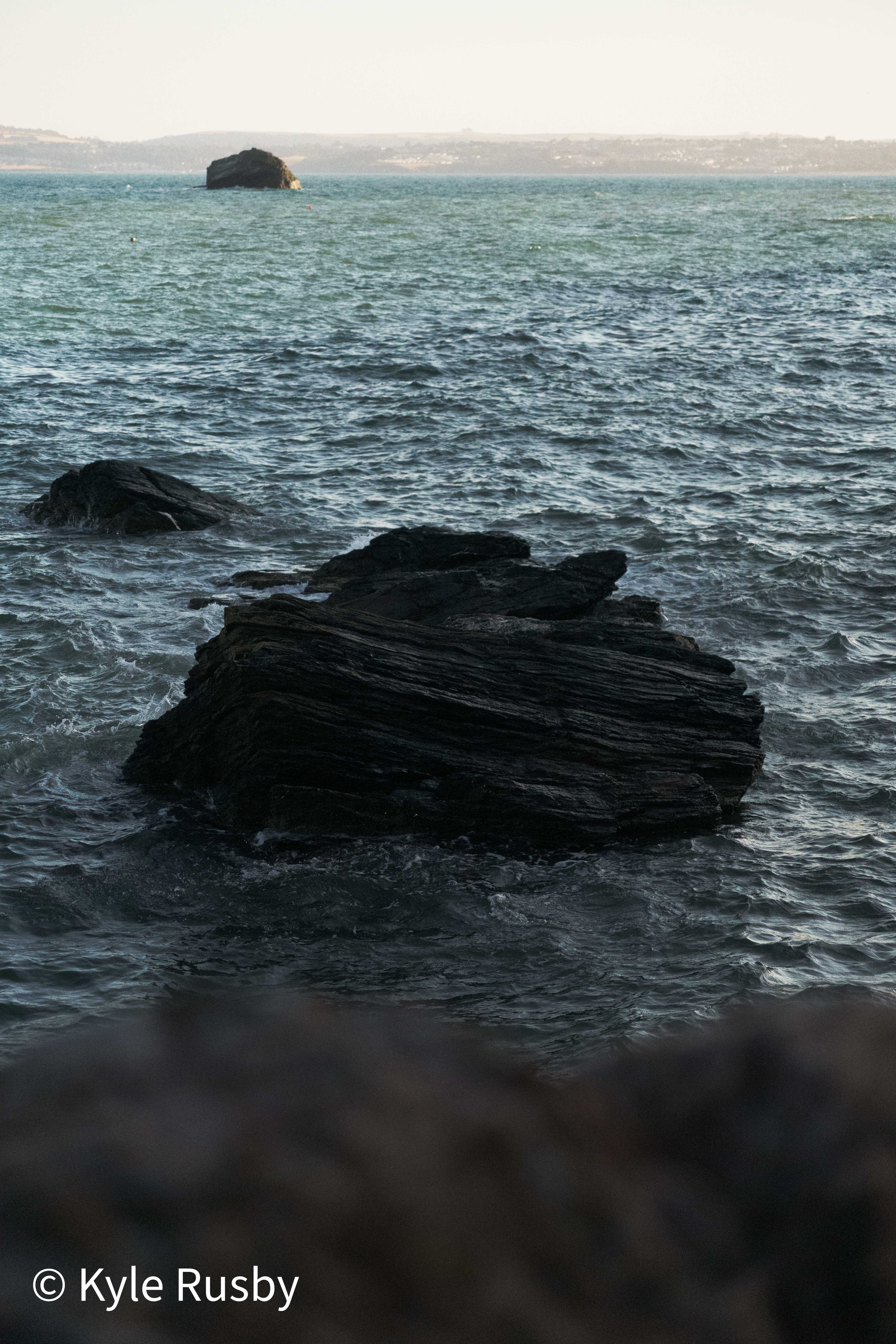 A dark layered rock breaks the surface of deep blue-grey water, a rock islet small on the pale horizon
