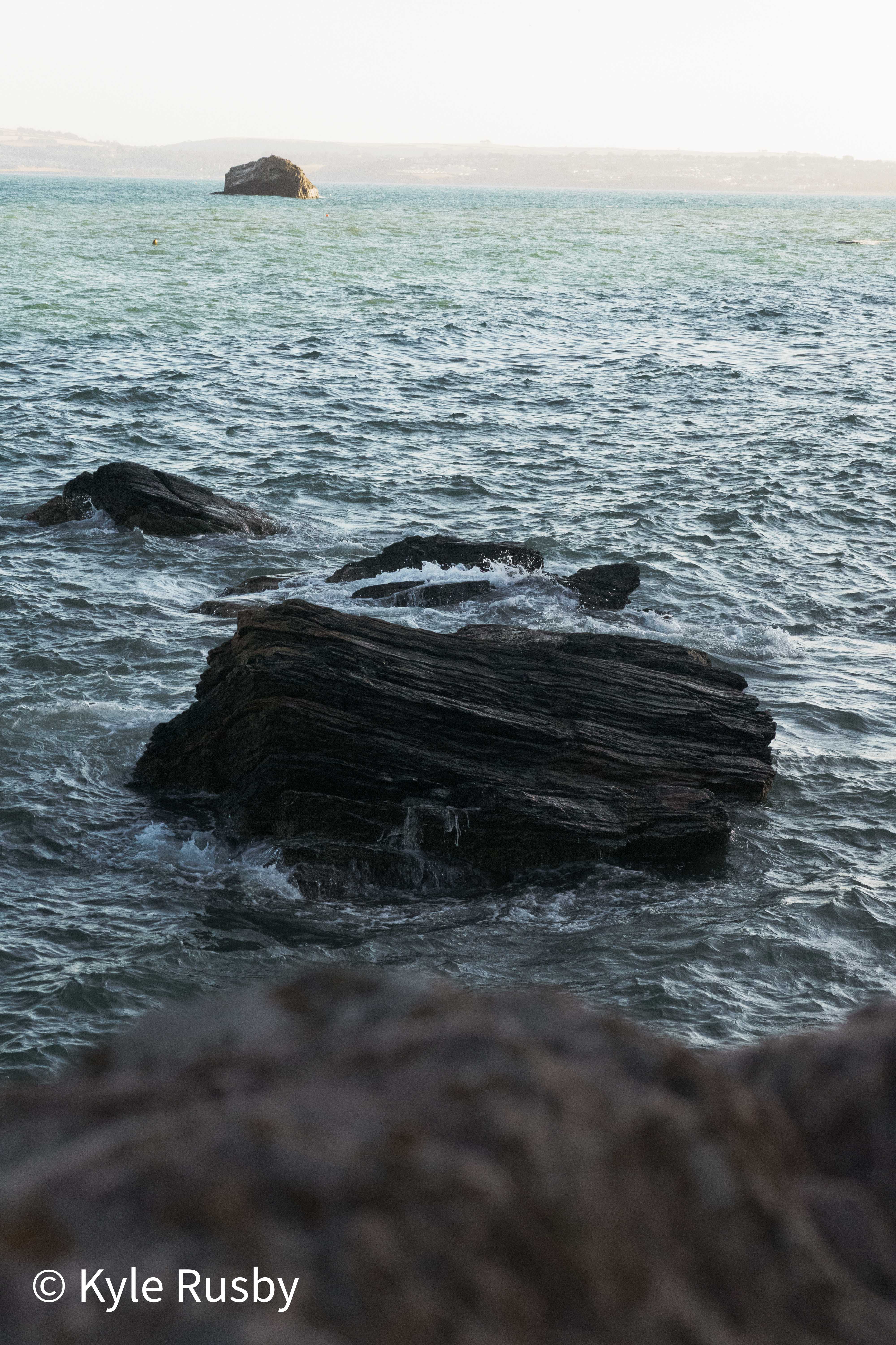 The same dark rocks from higher up, white water working through the channels between them towards the islet