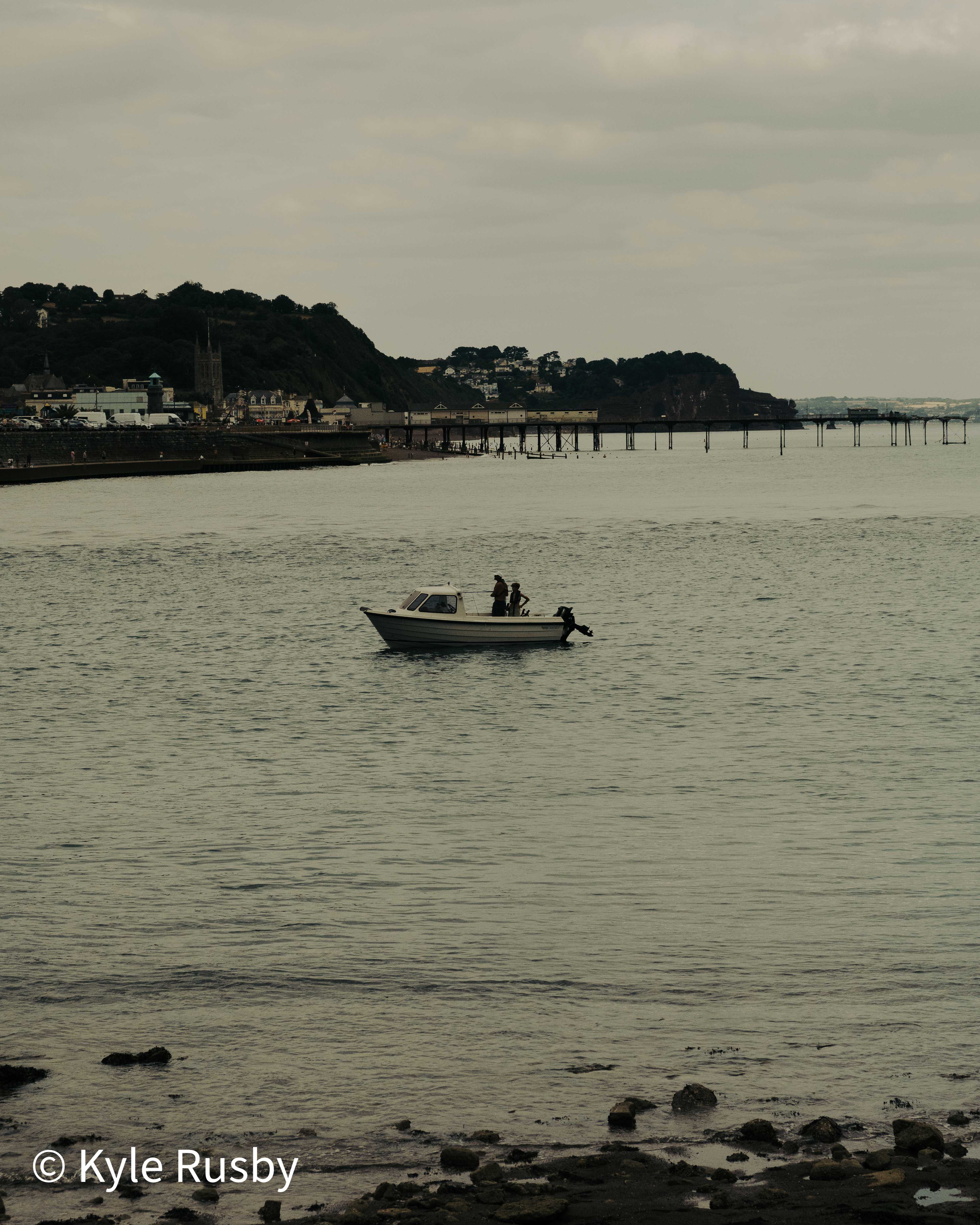 Two figures stand in a small open boat on flat grey water, the seafront town, its church tower and long pier stretched out behind them