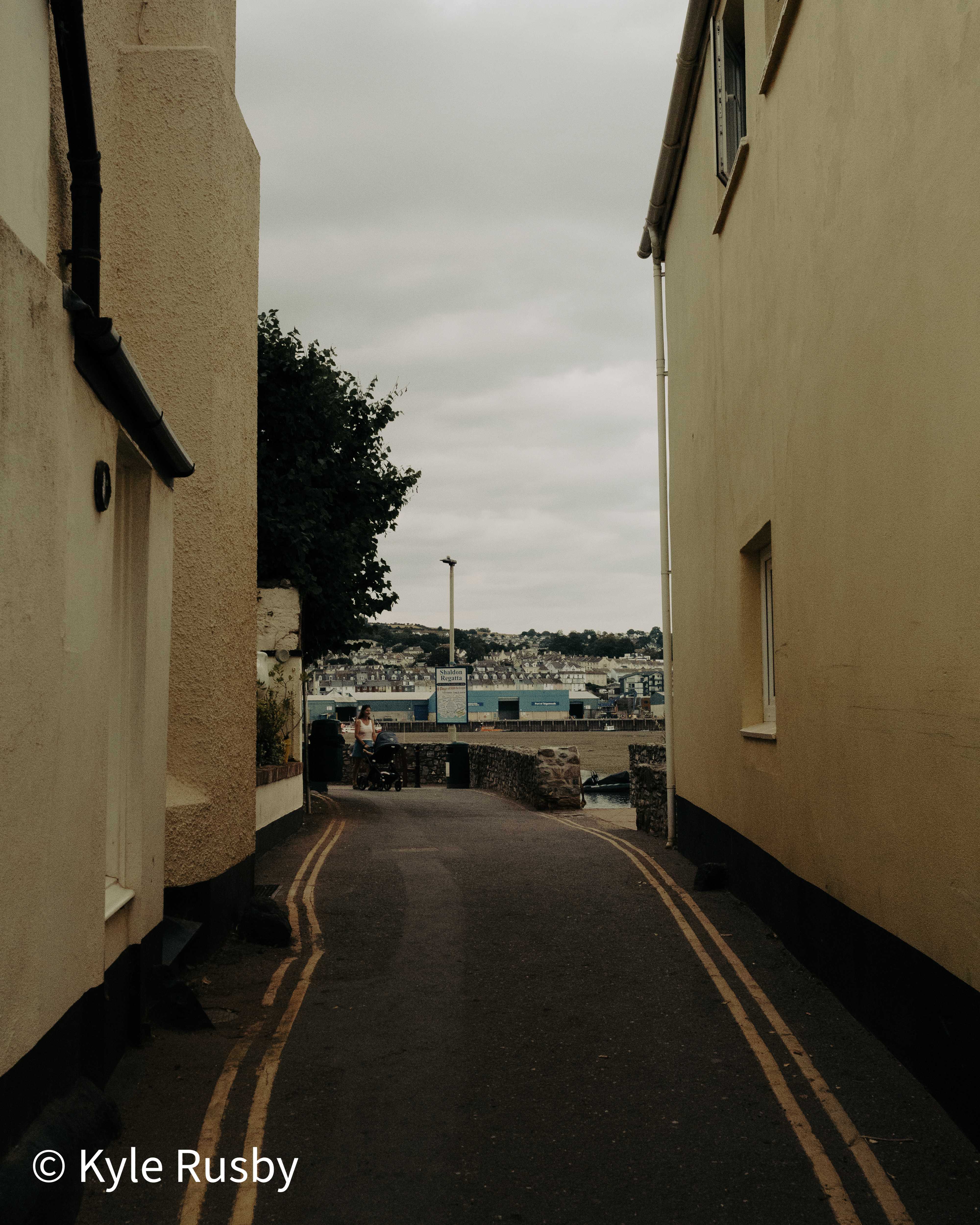 A narrow lane between tall rendered walls runs down towards the estuary, double yellow lines curving away to a blue regatta hut and the town rising beyond
