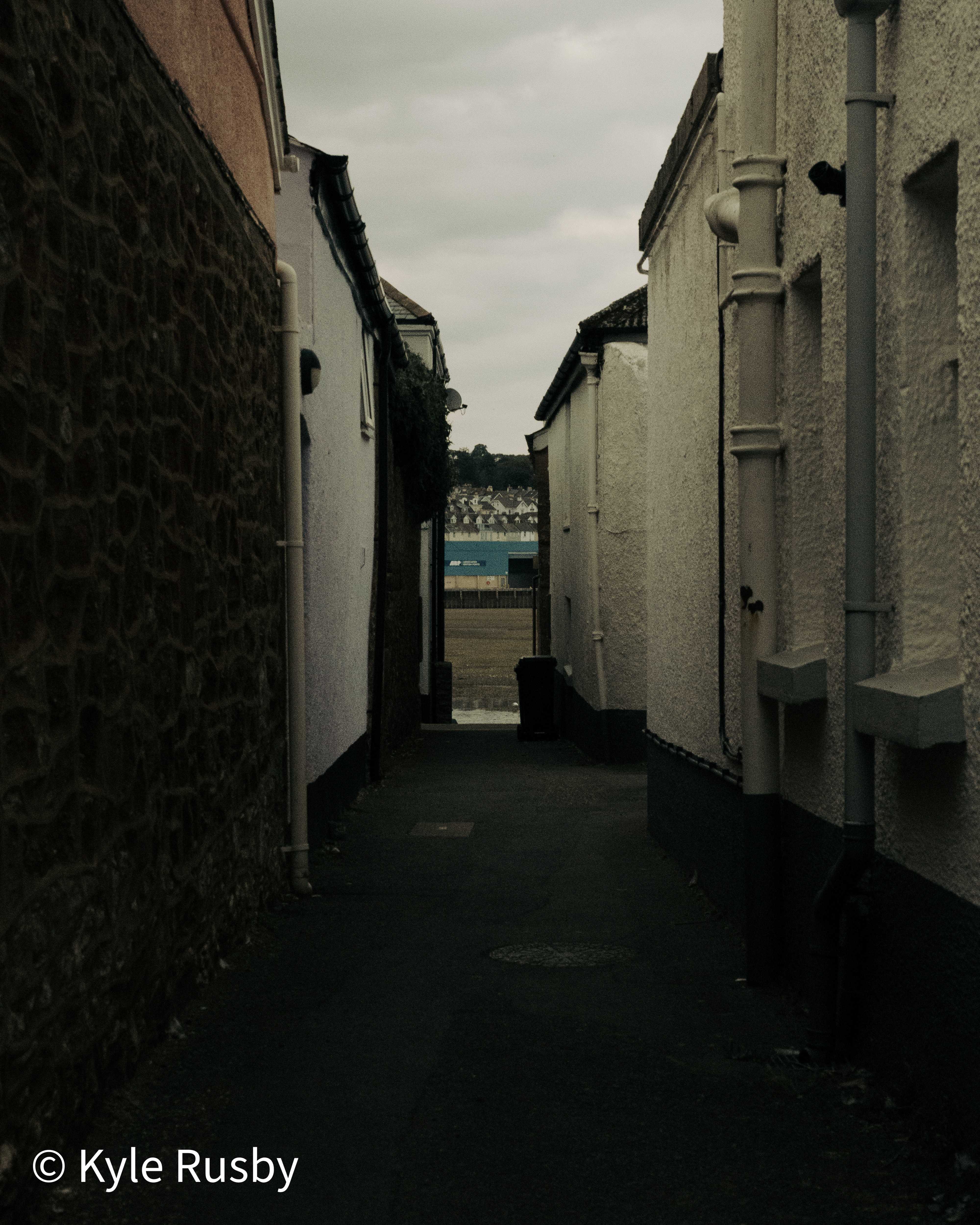 A dark, narrow lane runs between a rubble stone wall and white rendered houses, opening at the far end onto the bright estuary and the town beyond