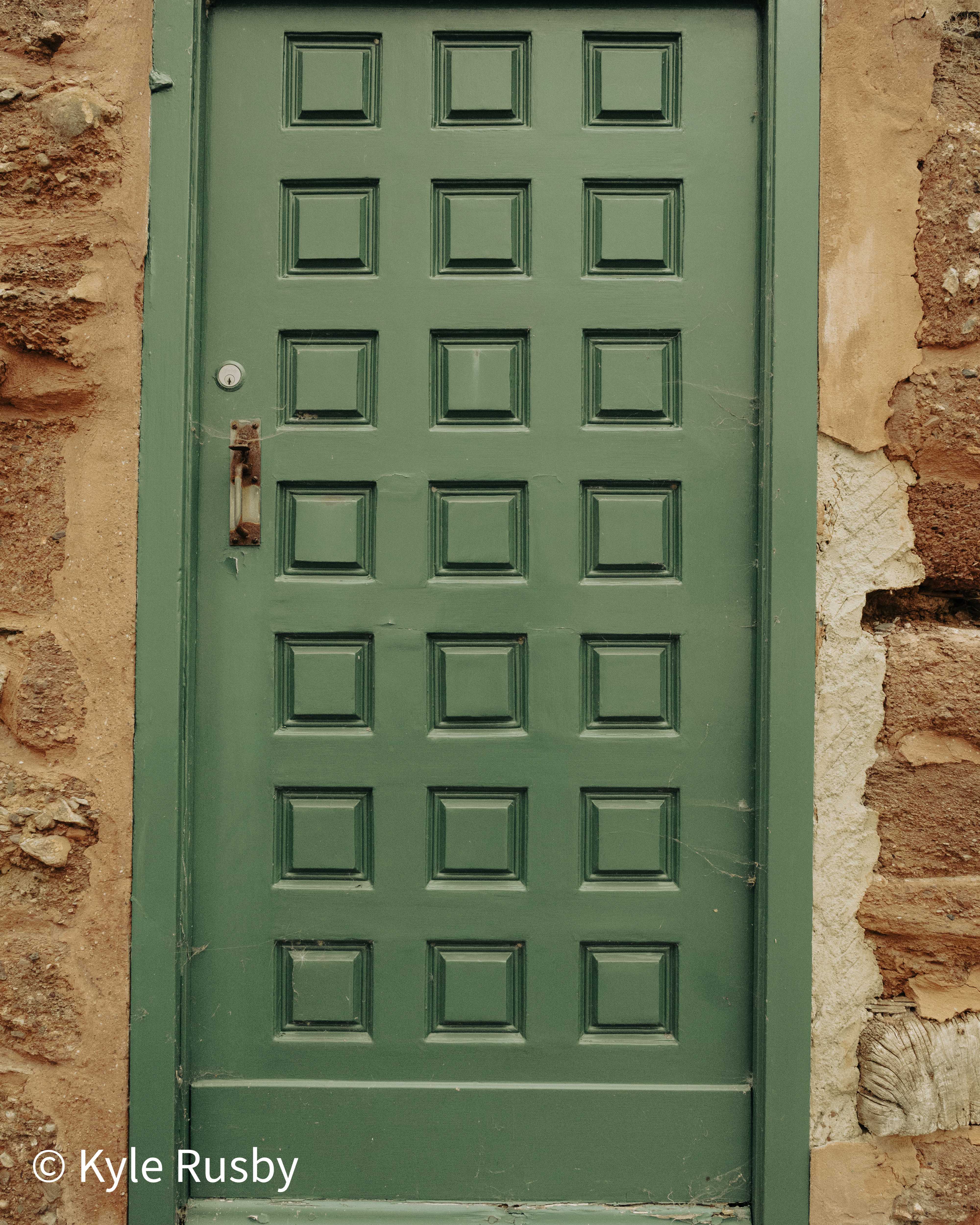 A heavily panelled green door with a worn brass letterplate set into a rough red sandstone and lime-mortar wall