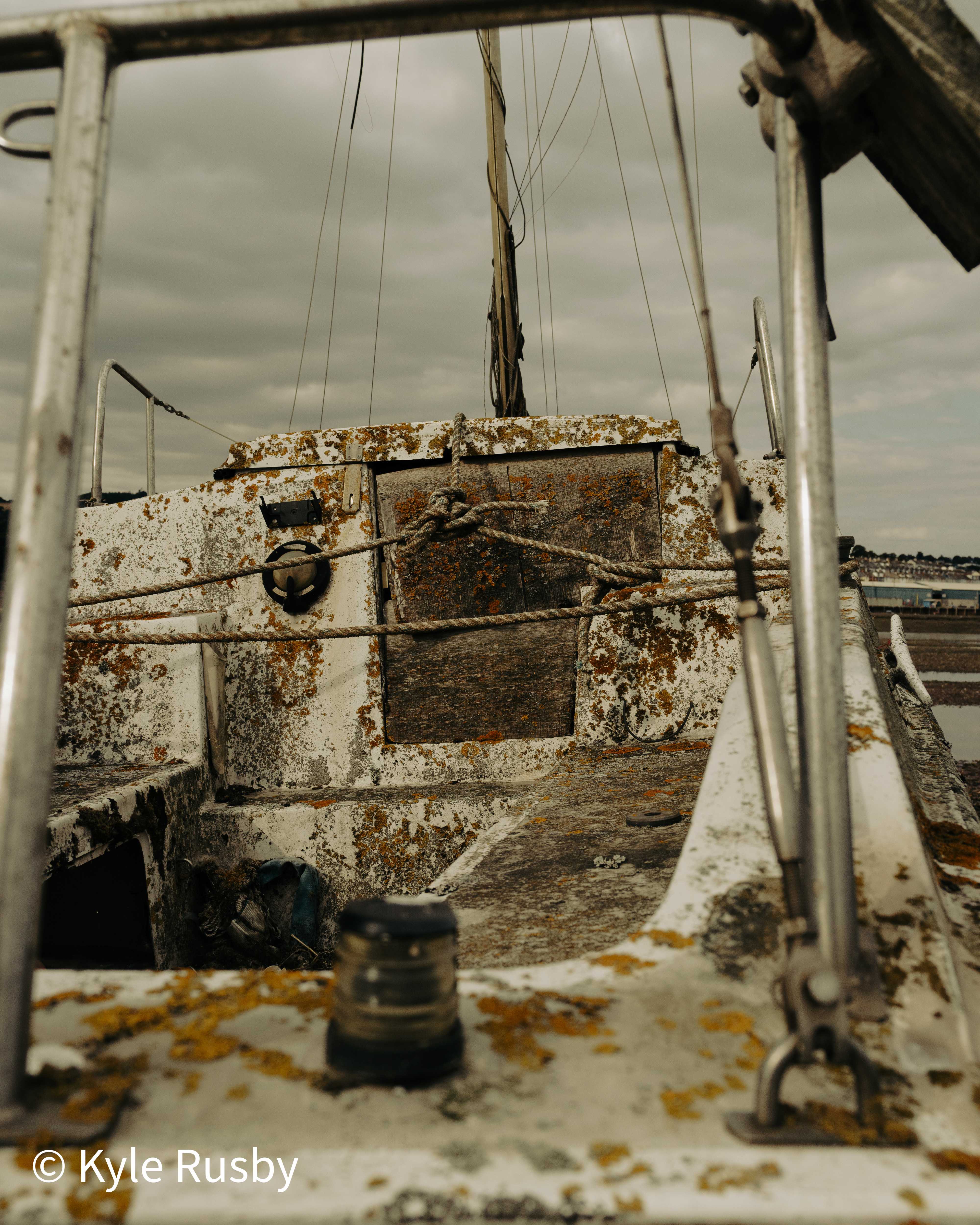 A glass and brass riding light sits on the lichen-spattered foredeck of a derelict boat, framed by steel guard rails, the estuary and far shore beyond
