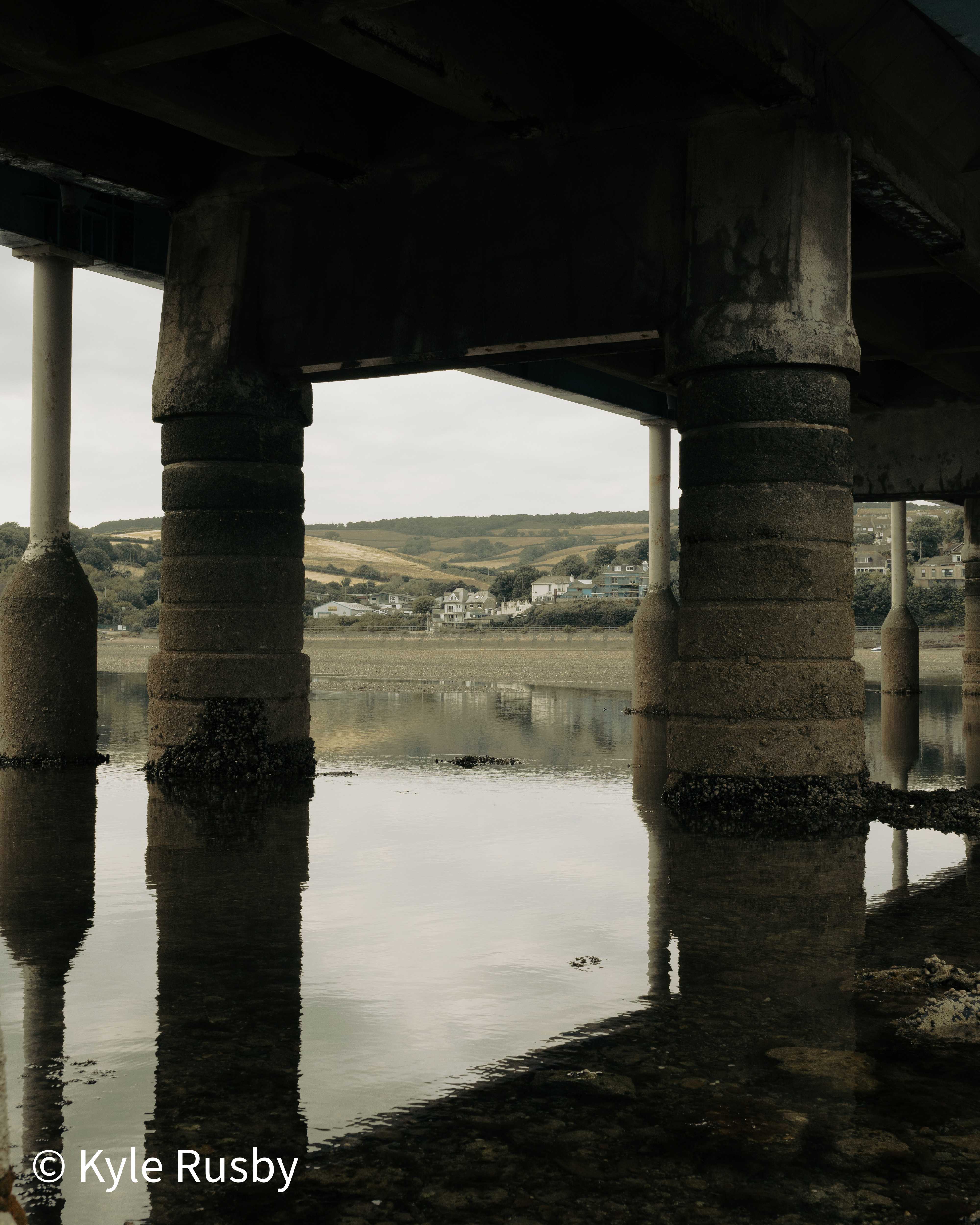 Barnacled concrete piers hold the underside of a road bridge above still estuary water, a gap between them framing hills and white houses on the far bank