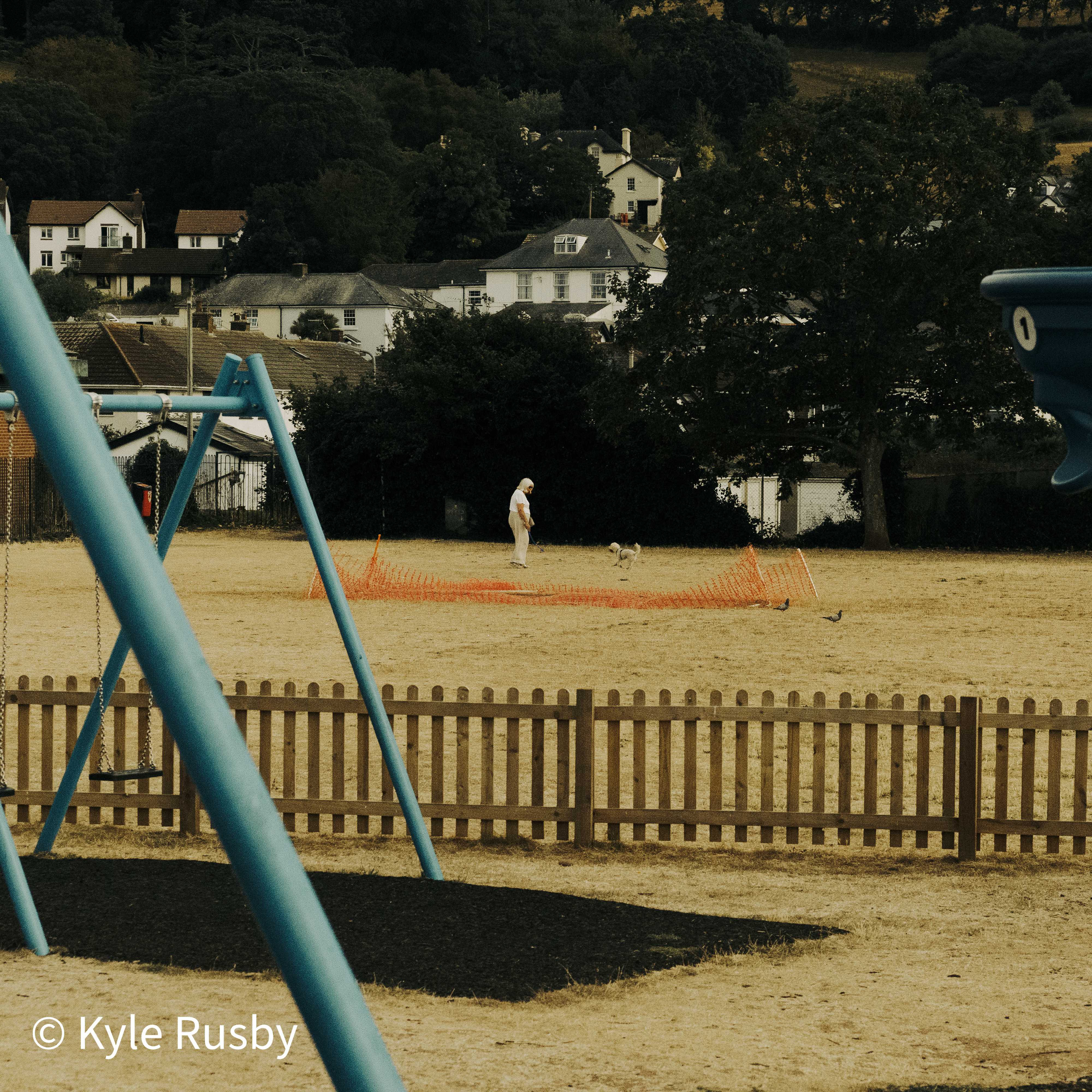 Blue swing frames stand in the foreground of a burnt-dry recreation ground, a man and a small white dog beyond an orange safety fence, houses climbing the wooded hill behind