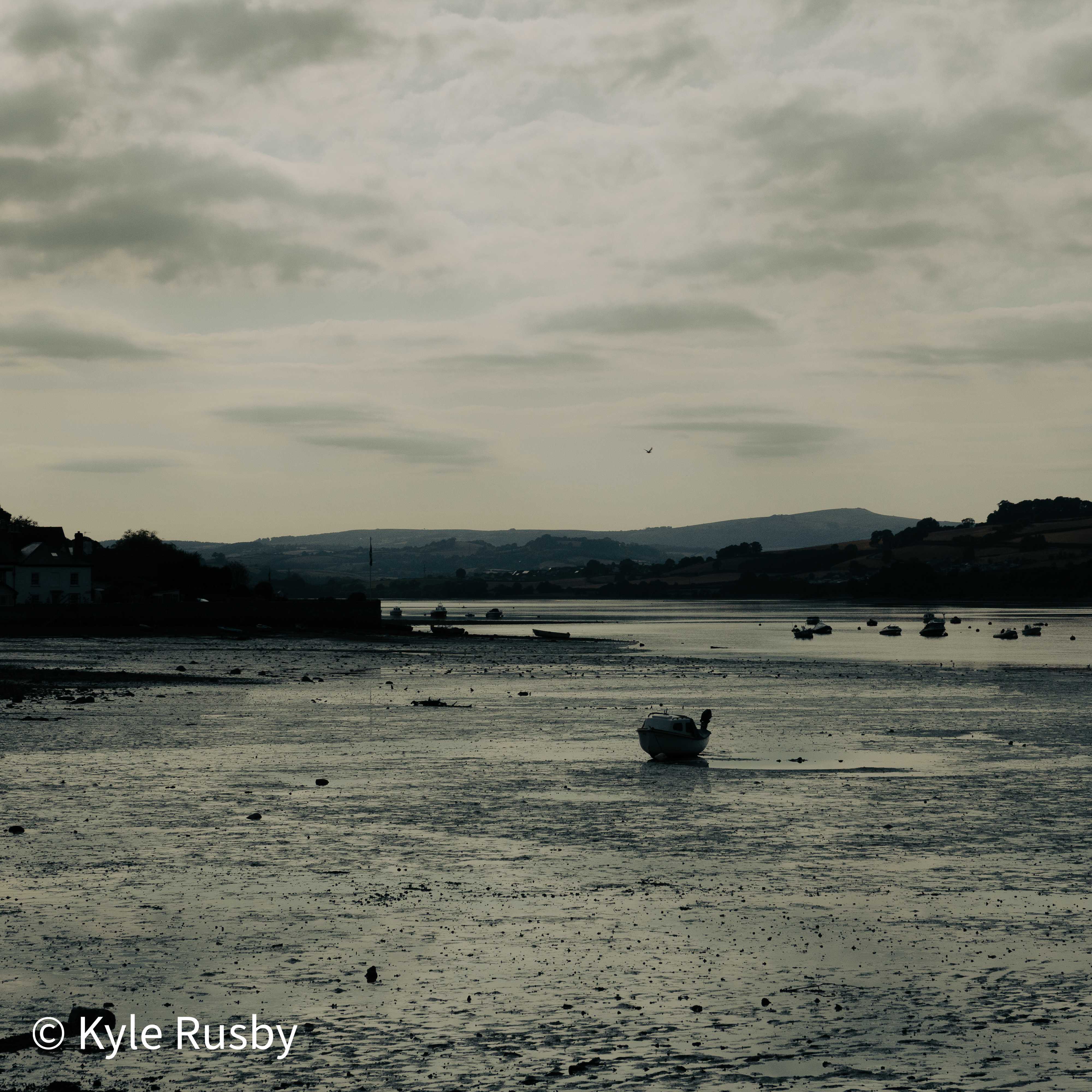 A single small boat sits aground on wide glistening mud, the rest of the fleet moored far off and moorland hills rising pale beyond