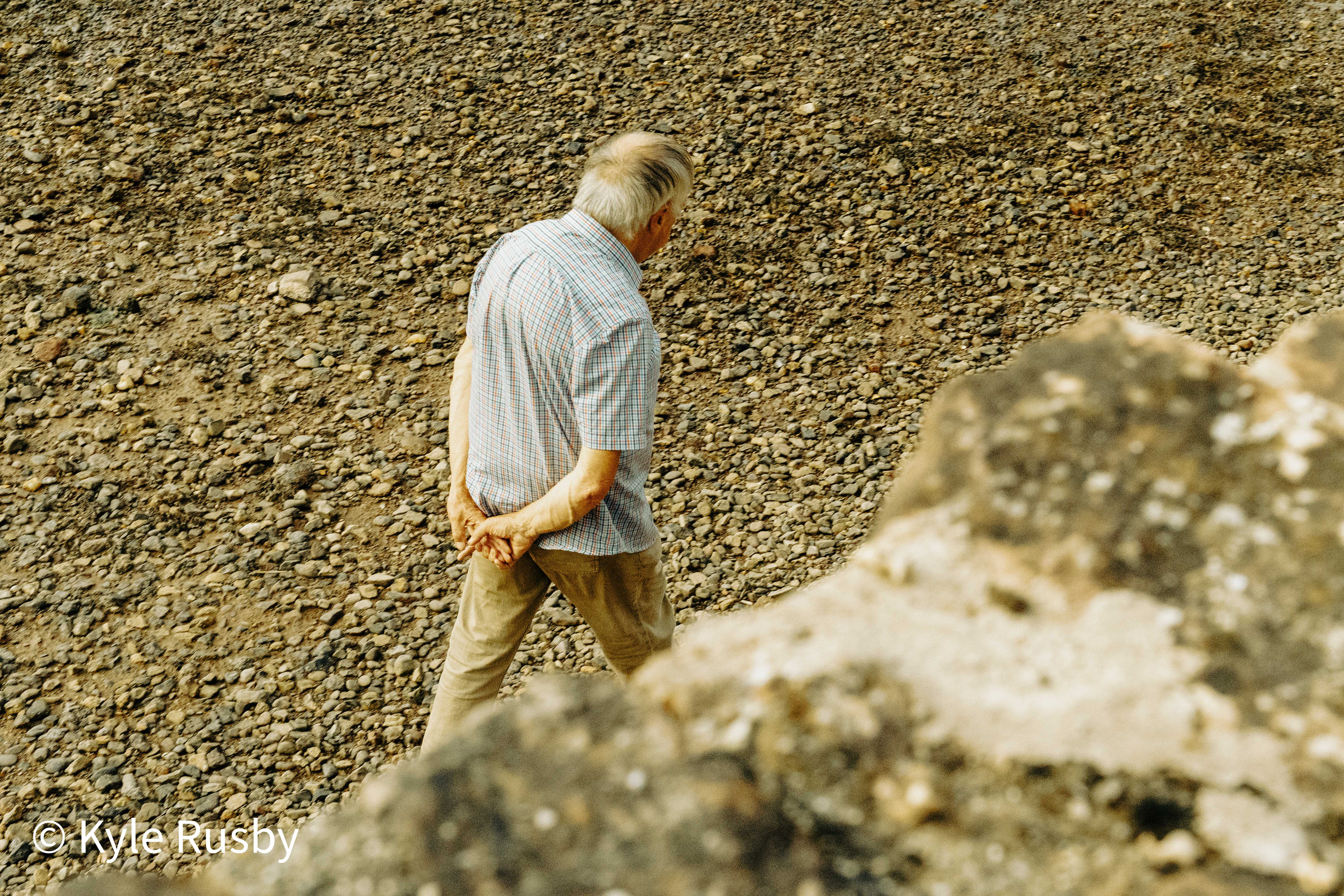 An older man in a checked shirt walks head-down across dry shingle with his hands clasped behind his back, a pale boulder blurred across the foreground