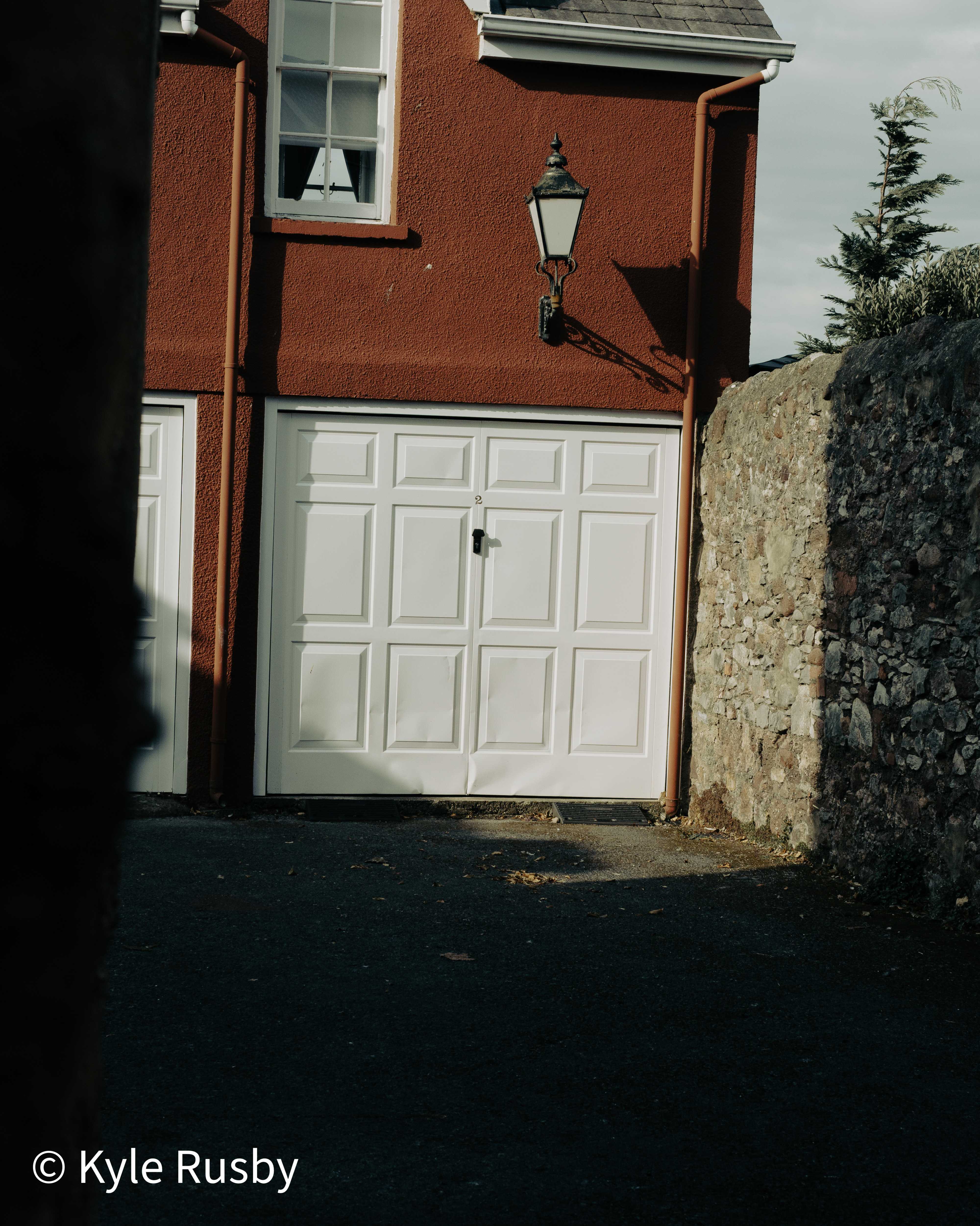 A black carriage lantern throws its shadow across the red rendered wall of a house above a white panelled garage door, framed by a dark tree trunk and a rubble stone wall