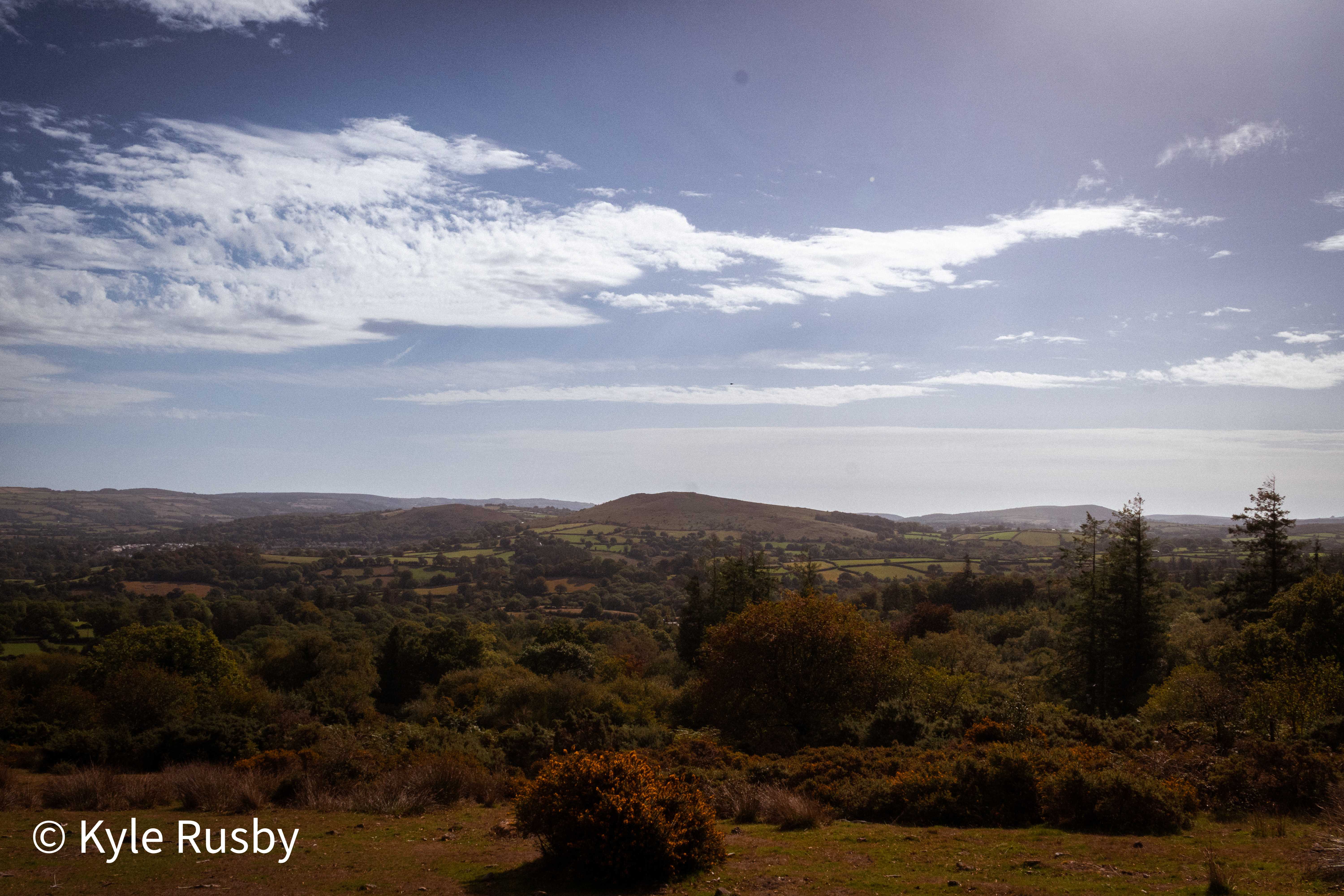 A wide view from the edge of the moor over wooded valley and hedged fields to distant hills, under a big sky of high broken cloud